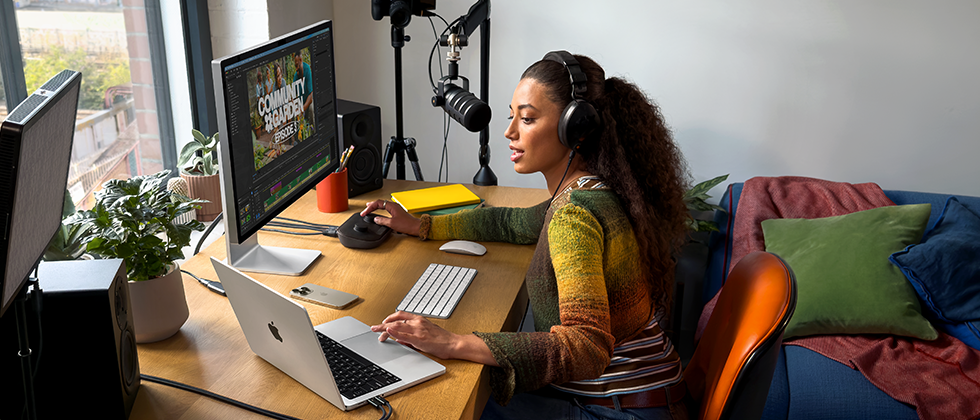 امرأة في منزلها تعمل على مكتبها مع توصيل MacBook Pro بشاشتين خارجيتين، ترتدي سماعات رأس وتتحدث في ميكروفون