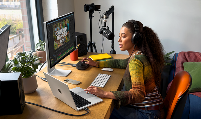 امرأة في منزلها تعمل على مكتبها مع توصيل MacBook Pro بشاشتين خارجيتين، ترتدي سماعات رأس وتتحدث في ميكروفون