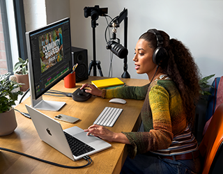 امرأة في منزلها تعمل على مكتبها مع توصيل MacBook Pro بشاشتين خارجيتين، ترتدي سماعات رأس وتتحدث في ميكروفون